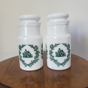 Pode incluir: Dois frascos de cerâmica branca com desenhos de folhas verdes e uma tampa branca. Os frascos estão sobre uma mesa de madeira.