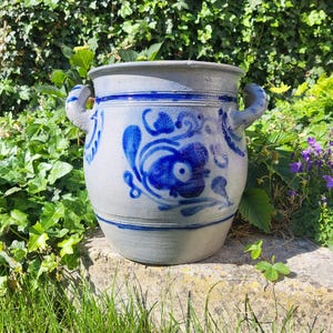 Può includere: Un vaso di ceramica grigia con disegni floreali blu e due manici. Il vaso ha un'ampia apertura ed è decorato con fiori e volute blu. Il vaso è appoggiato su una superficie di pietra, con vegetazione sullo sfondo.