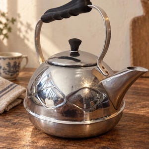 May include: A polished silver-tone metal tea kettle with a rounded body featuring embossed diamond-shaped patterns. It has a black ergonomic handle, a matching black knob on the removable lid, and a wide, angled spout for pouring hot water.