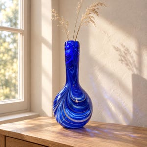 May include: A tall, bulbous glass vase featuring a vibrant cobalt blue base with swirling white and translucent streaks throughout the body. The vase has a narrow neck and contains two thin, dried beige stalks of ornamental grass.