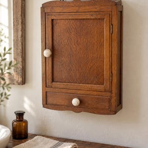 May include: A wall-mounted wooden medicine cabinet featuring a rectangular frame with a single hinged door and a bottom drawer. The cabinet is constructed from medium-brown wood with visible grain and includes two round, off-white ceramic knobs.