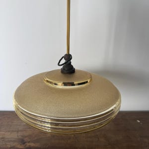 May include: A gold-colored pendant light with a round, textured glass shade. The shade has three horizontal lines around the bottom.