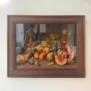 Puede incluir: Pintura enmarcada de naturaleza muerta con diversas frutas, como uvas, melocotones, peras y sandía. La obra de arte está ambientada sobre un fondo oscuro y enmarcada en un marco de madera oscura y ornamentada.