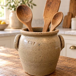 Op de afbeelding: Een beige keramieken pot met twee handvatten aan de zijkanten en de tekst 12J in reliëf op de voorzijde. Drie houten keukengerei, waaronder twee lepels en een platte spatel, staan in de pot en steken naar boven uit.