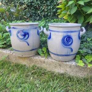 Puede incluir: Dos macetas de cerámica gris con diseños azules en espiral y línea. Cada maceta tiene dos asas y se asienta sobre una repisa de hormigón contra un fondo de follaje verde. Adecuado para exteriores.