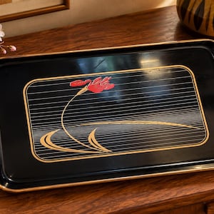 Puede incluir: Una bandeja de servicio rectangular negra con esquinas redondeadas y un borde dorado. El centro presenta un diseño gráfico de una flor roja con un tallo largo, sobre un fondo de finas líneas horizontales doradas y un borde dorado.