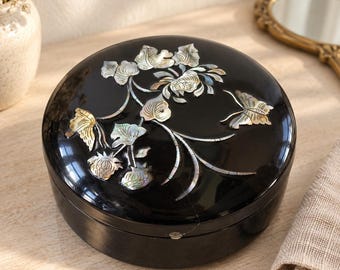 May include: A round black lacquer box featuring an iridescent mother-of-pearl inlay on the lid. The inlay depicts a floral branch with multiple leaves and blossoms, accompanied by a butterfly motif with shimmering, multi-colored accents.