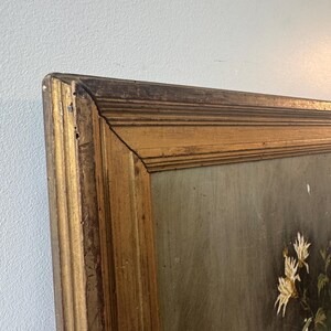 Op de afbeelding: Hoekweergave van een ingelijst schilderij met een goudkleurige houten lijst. Het schilderij toont witte bloemen met groene bladeren tegen een gedempte achtergrond. De lijst heeft een antieke uitstraling.