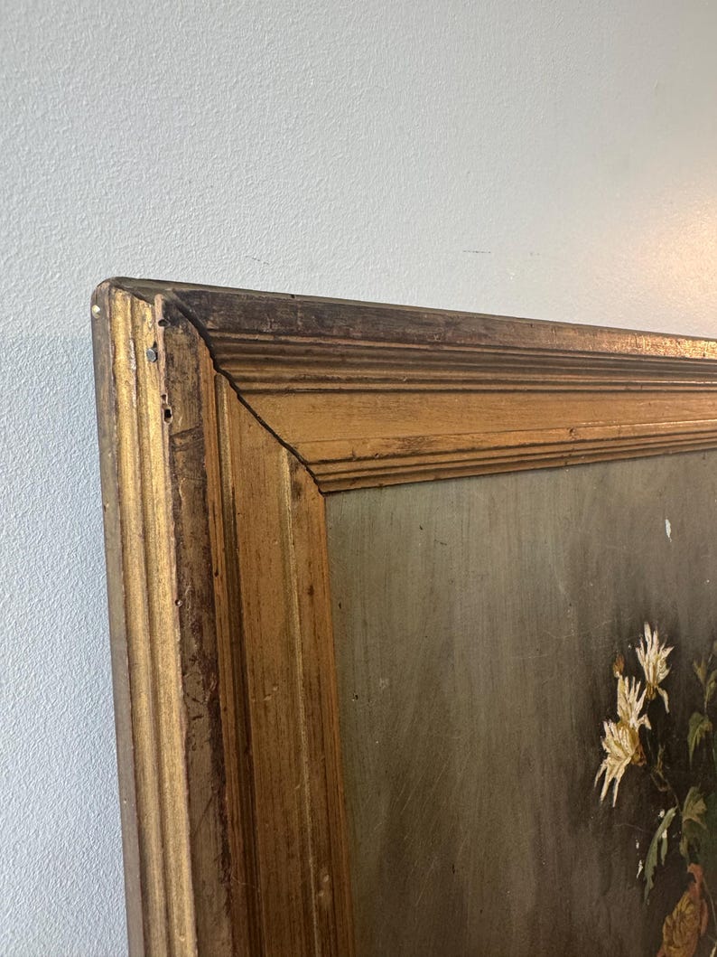 Op de afbeelding: Hoekweergave van een ingelijst schilderij met een goudkleurige houten lijst. Het schilderij toont witte bloemen met groene bladeren tegen een gedempte achtergrond. De lijst heeft een antieke uitstraling.