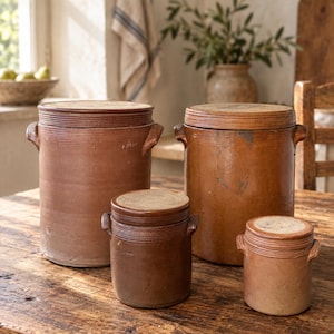 May include: A set of four cylindrical brown stoneware crocks with matching lids and side handles. The collection includes two large crocks and two smaller crocks, all featuring a smooth, earthy finish and a consistent, functional design.