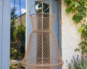 Puede incluir: Una cesta colgante de alambre con una estructura de dos niveles en forma de campana hecha de malla metálica de color marrón oxidado. La cesta presenta una abertura circular con un asa de alambre negro y una cuerda central vertical.