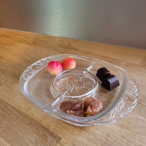 Puede incluir: Un plato de servir de vidrio transparente con cuatro compartimentos. El plato contiene dos caramelos rosas, dos caramelos marrones y dos cuadrados de chocolate negro.