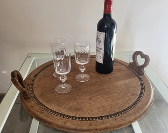 May include: A round wooden serving tray with carved edges and heart-shaped handles. Three clear glass wine glasses and a bottle of red wine are on the tray.