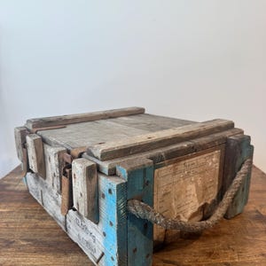 Pode incluir: Uma caixa de madeira desgastada com detalhes pintados em azul nos cantos, fechos de metal e uma alça de corda grossa na lateral. O exterior exibe texto impresso incluindo 13.500 Kg e 0.030m3, juntamente com uma etiqueta de papel desbotada.