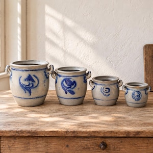 Può includere: Un set di quattro vasi in gres graduati con finitura grigio chiaro e motivi floreali dipinti a mano in blu. Ogni vaso presenta un corpo cilindrico, un bordo largo e due piccole maniglie laterali, con un design coerente in tutte le misure.