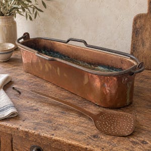 May include: An elongated rectangular copper basin with a dark metal rim and handle, featuring a weathered patina. Beside it lies a long-handled copper skimmer with a perforated flat head, showcasing a matching metallic finish and texture.
