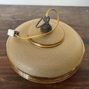 May include: A round, frosted glass ceiling light fixture with a gold trim and a yellow cord. The light fixture has a simple, vintage design.