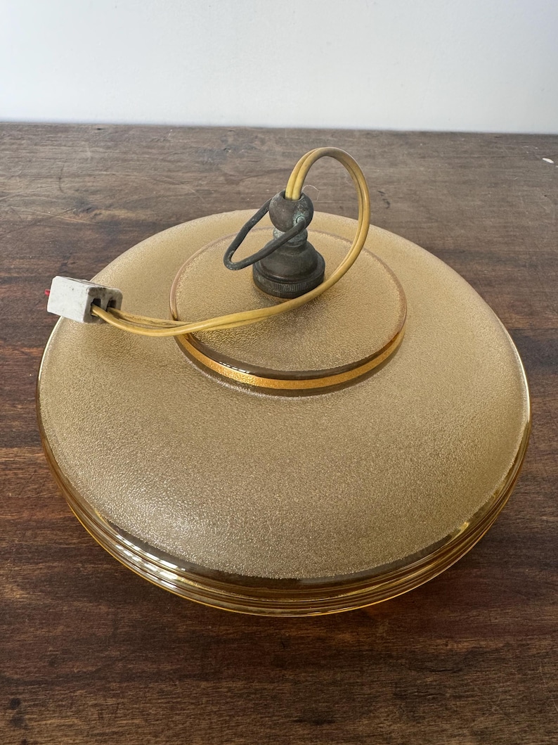 May include: A round, frosted glass ceiling light fixture with a gold trim and a yellow cord. The light fixture has a simple, vintage design.
