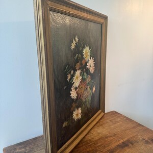Op de afbeelding: Een antiek bloemenschilderij in een goudkleurige houten lijst. Het kunstwerk toont een boeket witte en roze bloemen met groen blad tegen een donkere achtergrond. Het schilderij wordt getoond op een houten oppervlak.