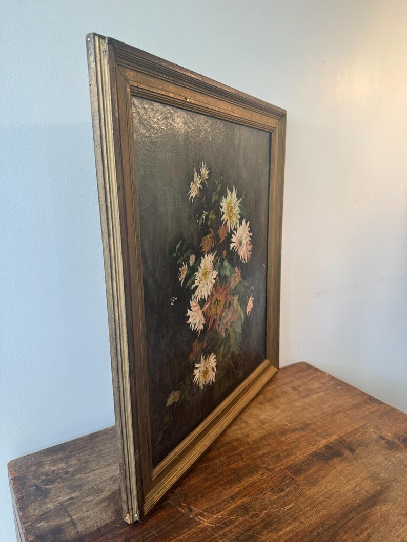 Op de afbeelding: Een antiek bloemenschilderij in een goudkleurige houten lijst. Het kunstwerk toont een boeket witte en roze bloemen met groen blad tegen een donkere achtergrond. Het schilderij wordt getoond op een houten oppervlak.