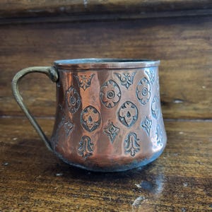 Puede incluir: Una taza de cobre con un diseño en relieve adornado. La taza tiene un asa de latón y un cuerpo redondeado.