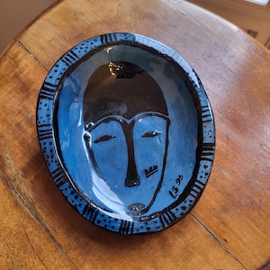 Könnte beinhalten: Eine blaue Keramik-Schale mit einem schwarz-weiß bemalten Gesichtsdesign. Die Schale hat einen schwarzen Rand mit weißen Punkten und Linien. Das Gesicht hat eine schwarze Krone und die Signatur "LS 93" ist auf der rechten Seite sichtbar.