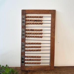 May include: A rectangular wooden abacus featuring twelve horizontal metal wires, each strung with ten natural brown wooden beads. The left side of the frame displays vertical black labels with the white printed numbers 10, 100, and 1000.