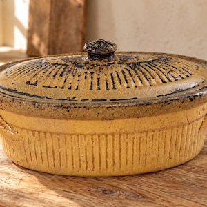 Peut inclure: Un plat à four ovale en céramique avec un corps beige texturé à stries verticales et deux poignées latérales. Le couvercle assorti présente un motif rayonnant brun foncé et un bouton central, avec une finition émaillée rustique.