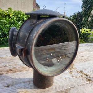 Op de afbeelding: Een vintage, ronde, zwarte metalen autokoplamp met een glazen lens. De koplamp heeft een metalen beugel aan de zijkant en een metalen dop bovenop.