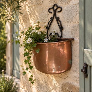 May include: A semi-circular copper wall planter featuring a black wrought iron scrollwork backplate. The planter is filled with green ivy vines and small white flowers, creating a contrast between the metallic copper finish and the foliage.