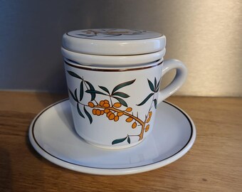 Puede incluir: Taza y platillo de té de cerámica blanca con un diseño floral verde y naranja. La taza tiene una tapa.