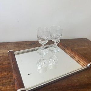 Op de afbeelding: Een spiegelende serveerschaal met een zilveren frame en houten handvatten, met twee heldere kristallen wijnglazen. De glazen worden weerspiegeld in de spiegel. De schaal staat op een houten oppervlak.