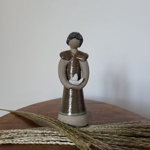 Peut inclure: Une figurine en céramique d'une personne tenant un oiseau blanc dans ses mains. La figurine est brune et verte avec une base blanche.