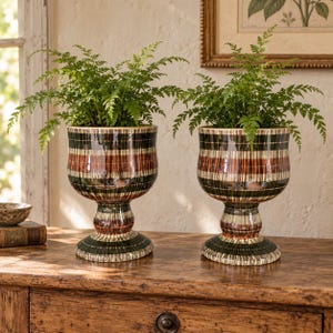 Può includere: Due vasi da fiori in ceramica su piedistallo abbinati con finitura lucida e strisce verticali nei toni del verde scuro, marrone e crema. Ogni vaso ha una forma a coppa larga e arrotondata sostenuta da una base circolare spessa.