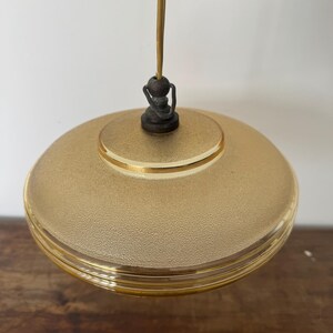 May include: A gold-toned glass pendant light with a textured surface and a gold trim. The light is suspended from a thin cord with a wooden bead at the top.
