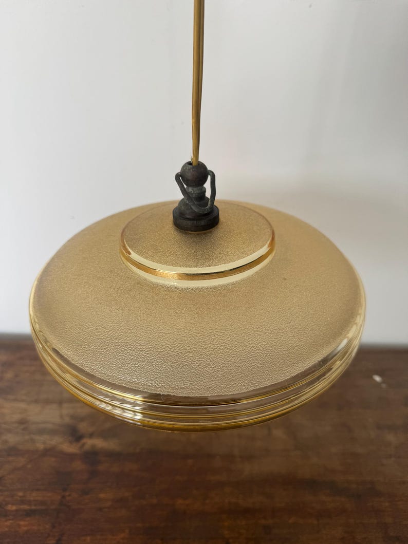 May include: A gold-toned glass pendant light with a textured surface and a gold trim. The light is suspended from a thin cord with a wooden bead at the top.