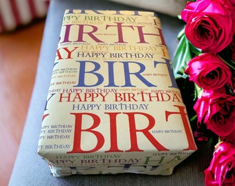 Op de afbeelding: Een ingepakt cadeau met een beige achtergrond en de woorden "Happy Birthday" in rood, blauw en paars. Het cadeau ligt op een grijze ondergrond, met een boeket rode rozen op de achtergrond.