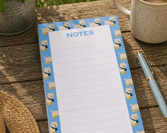 Op de afbeelding: Een rechthoekig notitieblok met een wit gelinieerd midden en het woord NOTES in blauw bovenaan gedrukt. De rand heeft een herhalend patroon van kleine witte honden met blauwe sjaals op een lichtblauwe achtergrond, verticaal geplaatst.