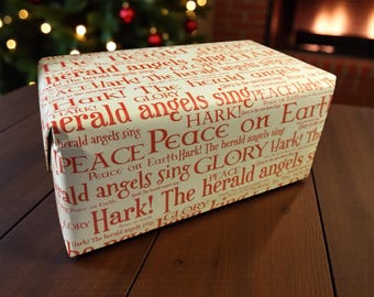 Op de afbeelding: Een ingepakte geschenkdoos met crèmekleurig papier met rode tekst met de tekst "Hark! The herald angels sing" en "Peace on Earth". Het cadeau staat op een houten oppervlak, met een kerstboom en een open haard op de achtergrond.