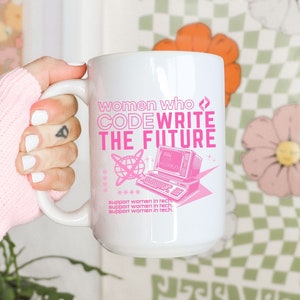 May include: White ceramic mug with a pink design that says "women who CODE WRITE THE FUTURE" with a retro computer graphic. The text also says "support women in tech" three times.