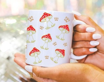 May include: White ceramic mug with a red and white mushroom pattern. The mushrooms are surrounded by small gold butterflies.