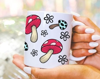 May include: White ceramic mug with a red and white mushroom design and blue and white mushroom design. The mug has a handle and is being held by a hand.