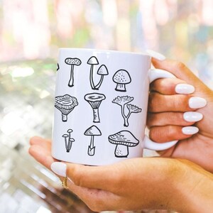 May include: White ceramic mug with a black and white illustration of various mushrooms. The mug has a handle on the right side.
