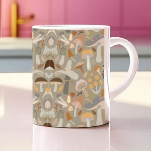 May include: A white ceramic mug with a handle features a pattern of colorful mushrooms and leaves on a light gray background.