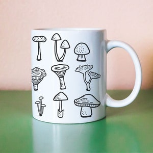 May include: White ceramic mug with a black outline drawing of various mushrooms. The mug has a handle on the right side.