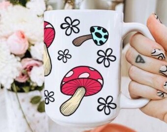 May include: White ceramic mug with a colorful mushroom and flower design. The mushrooms are red, blue, and yellow with white polka dots. The flowers are white with black outlines.