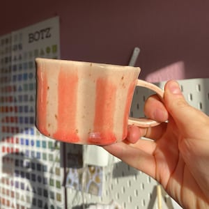 Könnte beinhalten: Eine handgefertigte Keramiktasse mit vertikalen Streifen in Koralle und Creme. Die Tasse hat eine abgerundete Form mit einem Henkel und wird vor einem rosa Hintergrund gehalten. Die Tasse wird wahrscheinlich zum Trinken von Kaffee oder Tee verwendet.