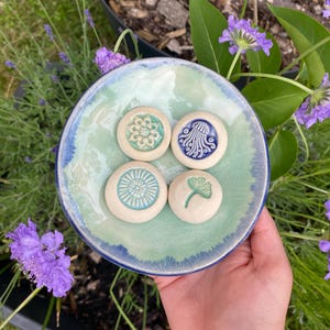 May include: A round ceramic dish with a light green and blue glazed finish containing four small, circular ceramic tokens. Each token features a distinct embossed design: a flower, a jellyfish, a sunburst pattern, and a ginkgo leaf.