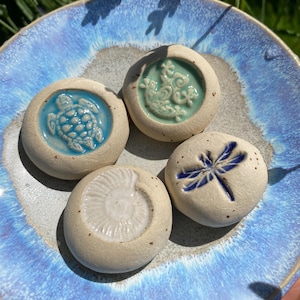 Könnte beinhalten: Vier kleine, runde Keramiksteine mit geprägten Designs einer Schildkröte, einer Libelle und anderer Meereskreaturen, präsentiert auf einem blauen Keramikteller. Die Steine sind cremefarben mit blauen und türkisfarbenen Akzenten. Der Teller hat eine blaue und graue Glasur.
