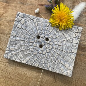 Könnte beinhalten: Ein rechteckiger, hellgrauer Keramik-Seifenhalter mit einem strukturierten, spitzenartigen Muster. Der Halter hat drei Abflusslöcher und ist mit einer gelben Pusteblume, lila Blumen und getrockneten Gräsern verziert. Auf einer Holzoberfläche.